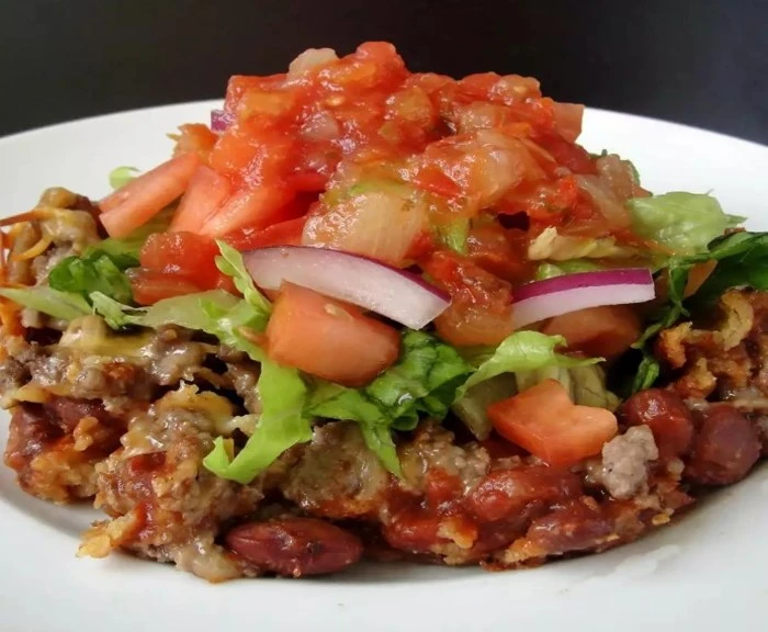 Ein frisch gebackener Taco-Auflauf in einer Auflaufform, garniert mit geschmolzenem Cheddar-Käse, gehackten Tomaten, Eisbergsalat, Salsa und einem Klecks Sauerrahm. Die goldbraunen Mais-Tortilla-Chips bilden die knusprige Basis, während das würzige vegetarische Chili für eine herzhafte Füllung sorgt.